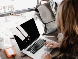 persoon met lang haar zit aan een bureau aan het raam, een rugzak naast haar, laptop voor haar neus
